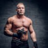 The brutal bearded muscular male holds dumbbell over grey vignette background.